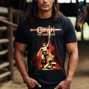 Könnte beinhalten: Schwarzes T-Shirt mit dem Filmtitel "Conan der Barbar" in gold- und weißfarbenen Buchstaben über einer Grafik der Filmfiguren. Die Grafik zeigt einen muskulösen Mann, der ein Schwert hält, und eine kniende Frau.