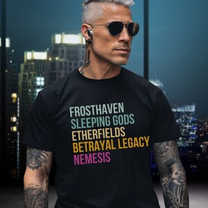 Może przedstawiać: Czarny T-shirt z grafiką w stylu vintage z napisami "Frosthaven", "Sleeping Gods", "Etherfields", "Betrayal Legacy" i "Nemesis" w różnych kolorach.
