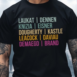 May include: Black t-shirt with a vintage-style graphic featuring a list of names: Laukat, Dennen, Knizia, Eisner, Dougherty, Kastle, Leacock, Daviau, Demaegd, and Brand.