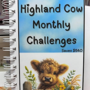 May include: A spiral-bound notebook with the title "Highland Cow Monthly Challenges" in black lettering against a blue sky background. The cover features a brown Highland cow illustration behind a wooden fence with yellow flowers. The text "Saves $540" is also present.