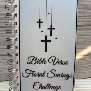 May include: A spiral-bound notebook with a white cover featuring black crosses and the text "Bible Verse Floral Savings Challenge." The notebook has a silver spiral binding and is displayed on a small black easel.
