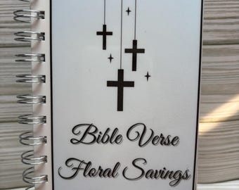 May include: A spiral-bound notebook with a white cover featuring black crosses and the text "Bible Verse Floral Savings Challenge." The notebook has a silver spiral binding and is displayed on a small black easel.