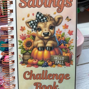 May include: A savings challenge book with a cute cartoon cow wearing a black and white checkered bow. The cow is sitting on a pumpkin with a sign that says "Hello Fall". The book cover is orange, yellow, and green with a white background. The text on the cover says "Savings Challenge Book" and "Saves $135+".