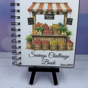 May include: A spiral-bound "Savings Challenge Book" with a "Farmer's Market" cover illustration. The cover art depicts a market stall with produce. The book is on a small black easel. The text "Saves $365" is visible.