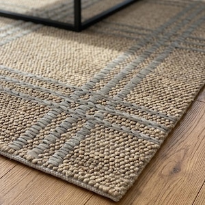 May include: A beige rectangular rug with a woven texture and a grey geometric pattern. The rug is on a wooden floor, with a black metal table leg in the background. The rug's design features intersecting lines.