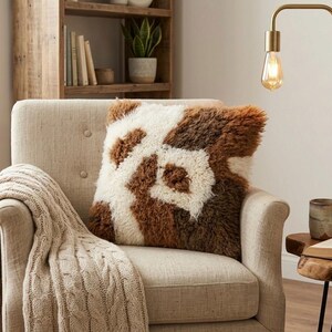 May include: A beige armchair with a decorative pillow featuring a brown and white pattern. A cable-knit throw blanket is draped over the arm. A gold lamp and a small wooden side table with a mug and book complete the cozy scene.