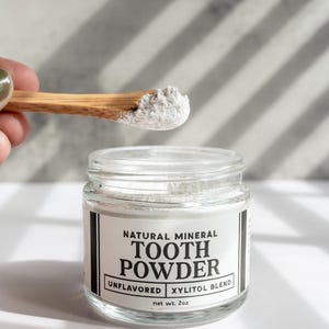 May include: A clear glass jar of natural mineral tooth powder with a wooden spoon. The label reads "TOOTH POWDER, UNFLAVORED, XYLITOL BLEND, net wt. 2oz." The spoon holds a small amount of the white powder.