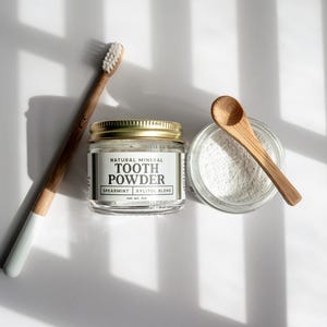 K&ouml;nnte beinhalten: Ein Set aus nat&uuml;rlichem Mineral-Zahnpulver, bestehend aus einer Bambus-Zahnb&uuml;rste, einem Glas mit Zahnpulver mit goldenem Deckel, einem kleinen Holzl&ouml;ffel und einem Glasbeh&auml;lter mit Pulver. Das Etikett des Glases lautet "Tooth Powder Spearmint Xylitol Blend".