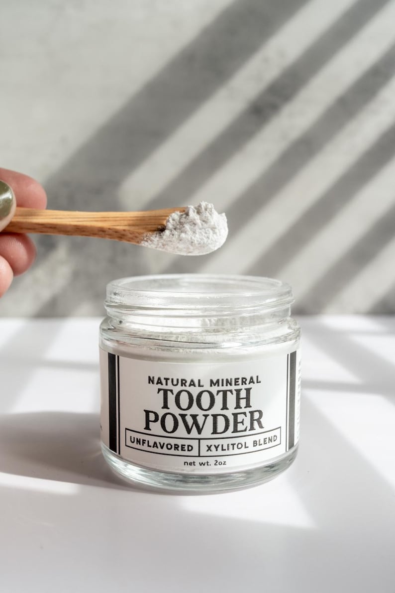May include: A clear glass jar of natural mineral tooth powder with a wooden spoon. The label reads "TOOTH POWDER, UNFLAVORED, XYLITOL BLEND, net wt. 2oz." The spoon holds a small amount of the white powder.