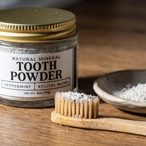 May include: A glass jar of natural mineral tooth powder with a gold lid, labeled with the text Natural Mineral Tooth Powder Peppermint Xylitol Blend net wt. 2oz (38g). A bamboo toothbrush with powder on the bristles and a small ceramic dish of powder.