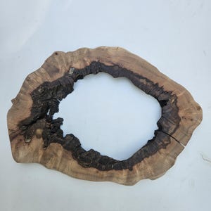 Op de afbeelding: Een natuurlijke houten plaat met een donkerbruin en lichtbruin kleurenschema. Het hout is in een cirkelvorm gesneden met een gat in het midden. Het hout heeft een ruwe textuur en zichtbare nerfpatronen.