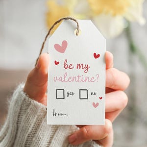 May include: A white gift tag with the text "be my valentine?" in red and pink script, with heart illustrations. The tag has boxes for "yes" and "no" and a space for "from:" and is held by a person.