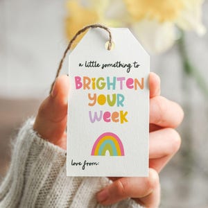 May include: A white gift tag with the text "a little something to BRIGHTEN YOUR WEEK" in colorful letters. A rainbow illustration is below the text, and the words "love from:" are at the bottom. The tag is held by a person.