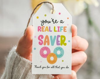 May include: A white rectangular gift tag with a brown string, featuring the text you're a REAL LIFE SAVER and Thank you for all that you do. The design includes colorful polka dots and three ring-shaped candy illustrations in pink, blue, and orange.