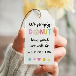 Pode incluir: Uma etiqueta de presente branca com o texto "We simply DONUT know what we will do WITHOUT YOU!" em preto, verde-azulado e amarelo. A palavra "DONUT" apresenta um gráfico de donut colorido. A etiqueta é segurada por uma pessoa.