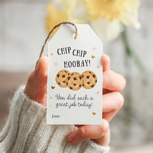 May include: A white gift tag with the text "CHIP CHIP HOORAY!" and "You did such a great job today!" features three watercolor chocolate chip cookies. The tag is held by a person wearing a gray sweater.