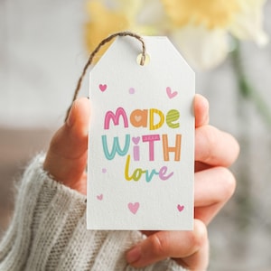 Pode incluir: Uma etiqueta de presente branca com as palavras "Made With Love" em letras coloridas e pequenos corações rosa. A etiqueta é segurada por uma pessoa que usa um suéter cinza. Uma corda marrom está presa à etiqueta.