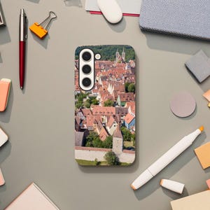 Può includere: Una custodia per telefono con una fotografia aerea colorata di una città europea con tetti di tegole rosse. La custodia ha un ritaglio dell'obiettivo della fotocamera nero con tre fori.