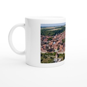 Könnte beinhalten: Eine weiße Keramiktasse mit einem bunten Bild einer europäischen Stadt mit roten Ziegeldächern und einer Steinmauer. Die Stadt ist von grünen Bäumen und blauem Himmel umgeben.