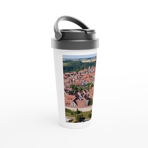Può includere: Tazza da viaggio bianca con coperchio e manico neri. La tazza presenta una foto di una piccola città con tetti in tegole rosse e una foresta verde sullo sfondo.