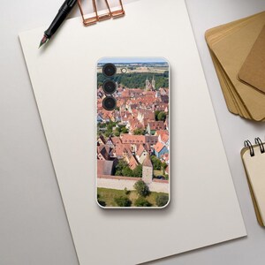 May include: A phone case featuring a colourful aerial photograph of a European town with red-tiled roofs and a tall tower. The case has a black camera bump with three lenses.