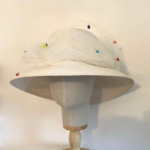 May include: A white hat with a wide brim and a white veil. The hat is decorated with colourful polka dots.