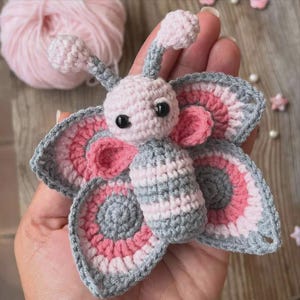 May include: A handmade crochet butterfly in shades of pink, gray, and white. The butterfly has large, detailed wings with a circular pattern, and a small body. The antennae and eyes are also visible. The butterfly is a craft item.