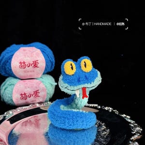 Puede incluir: Una serpiente de peluche azul hecha a mano con ojos amarillos y lengua roja está sobre una bandeja plateada. Dos ovillos de lana azul y verde con etiquetas rosas están en el fondo. Las etiquetas tienen caracteres chinos. También se ve el texto "@布丁| HANDMADE | 小红书".