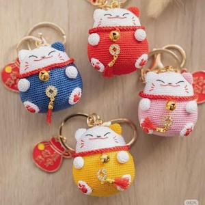 May include: A collection of handmade crochet cat keychains in various colors: red, blue, pink, and yellow. Each keychain features a smiling cat design with a gold bell and tassel. The keychains are attached to gold-colored key rings.