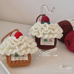Puede incluir: Dos monederos hechos a mano con forma de cupcake de ganchillo. Los monederos tienen una base marrón, glaseado blanco con volantes y una cereza roja encima. Cada monedero tiene una etiqueta que dice "Hand Made". Se ve hilo marrón en el fondo.