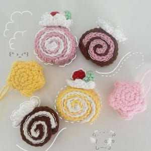 May include: A collection of handmade crochet food items, including pink, yellow, and brown cake rolls, a yellow star, and a yellow, star-shaped item. Each piece is detailed with white frosting and red and green accents.