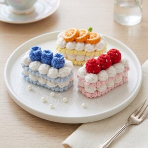 May include: Three rectangular crochet cakes on a white plate. One cake is light blue with blue berries, one is yellow with orange slices, and one is light pink with red berries. Each cake features white crocheted frosting dollops.