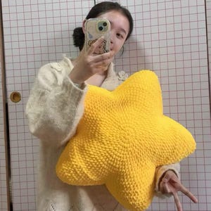 May include: A large, bright yellow star-shaped cushion. The cushion is made of a textured, knitted material. The background is a white wall with a grid pattern.