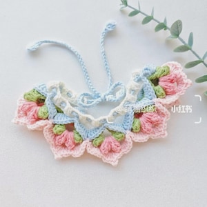 May include: A handmade crochet collar in pastel colors. The collar features a floral design with pink, green, and blue elements. It has a delicate, lacy appearance and is tied with a light blue string.