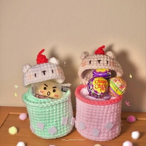 May include: Two handmade crochet containers, one mint green and one pink, each with a bear-shaped lid and a red pompom. The mint green container holds a snack, while the pink one contains lollipops. The containers are decorated with small white squares.