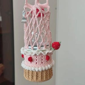 Puede incluir: Un portabotellas de agua de ganchillo rosa hecho a mano, diseñado para parecerse a un pastel. El soporte presenta una parte superior en forma de enrejado, un borde blanco decorativo y está adornado con acentos de fresas rojas. Un pequeño dije de tenedor plateado cuelga de la correa.