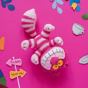 Puede incluir: Un juguete hecho a mano de ganchillo del Gato de Cheshire, a rayas rosas y blancas, con ojos amarillos y una gran sonrisa, sobre un fondo rosa brillante. Recortes de papel decorativos de hojas, una taza de té y letreros que dicen "Tea Party" y "Wrong Way" rodean al gato.