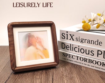 May include: A wooden picture frame with a light brown finish, displaying a blurry image of a person. The frame is sitting on a wooden table with a stack of books in the background. The books have white covers with black text that reads "six sigrande Delicious Pl SPECTORBOOK I saw its lights reflected in the sky and heard ther".
