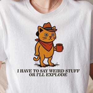 May include: White t-shirt featuring a cartoon cat wearing a cowboy hat and holding a mug. The cat is orange with a red bandana. The text on the shirt reads, "I HAVE TO SAY WEIRD STUFF OR I'LL EXPLODE."