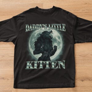 Pode incluir: Camiseta preta com um desenho de lobisomem rosnando em frente a uma lua cheia. O texto "DADDY'S LITTLE KITTEN" &eacute; impresso acima e abaixo da imagem em prata.