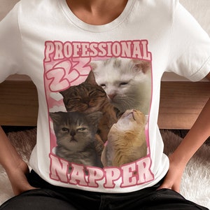 Op de afbeelding: Wit T-shirt met een roze afbeelding van vier katten en de tekst "Professional Napper".