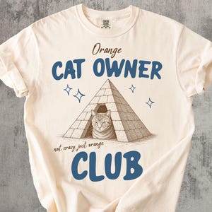 May include: Cream-coloured t-shirt with the text "Orange CAT OWNER CLUB" in blue and brown. The graphic features a cat inside a pyramid. The phrase "not crazy just orange" is also included.