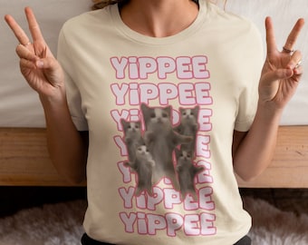 May include: A person wearing a cream colored t-shirt with the text "YIPPEE" repeated in pink letters. The shirt features a graphic of several dancing cats.
