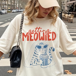 Könnte beinhalten: Eine beige Baseballkappe mit kleinem dunklem Logo und ein übergroßes cremefarbenes T-Shirt mit einer Grafik einer Katze, die eine Sonnenbrille und einen Schleier trägt und einen Kaffeebecher hält, mit dem rot-blauen Text "getting MEOWIED".
