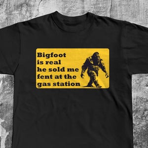 K&ouml;nnte beinhalten: Schwarzes T-Shirt mit einem gelben Schild, auf dem steht "Bigfoot is real he sold me fent at the gas station". Auf dem Schild ist eine Silhouette von Bigfoot abgebildet.