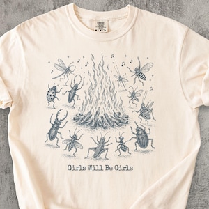 Peut inclure: Un t-shirt à manches courtes de couleur crème avec une illustration à l'encre bleu foncé représentant divers insectes dansant autour d'un feu de camp. Le texte "Girls Will Be Girls" est imprimé sous le graphique central du feu.