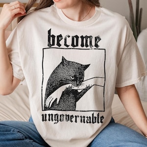 Op de afbeelding: Cr&egrave;mekleurig T-shirt met een zwarte kattenafbeelding in een zwart rechthoekig kader. De tekst "become" staat boven de kat en "ungovernable" eronder. De afbeelding toont een grote en een kleine kat met een hand.