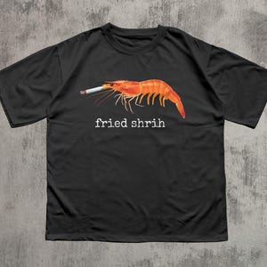 May include: Black t-shirt with a graphic of a red shrimp smoking a cigarette. The text "fried shrih" is printed below the shrimp in white letters. The shirt is made of a soft material.