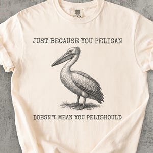 Puede incluir: Camiseta color crema con una ilustración de un pelícano y el texto "JUST BECAUSE YOU PELICAN DOESN'T MEAN YOU PELISHSHOULD". El gráfico es un dibujo detallado en blanco y negro de un pelícano.