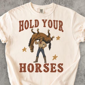 Op de afbeelding: Een crèmekleurig T-shirt met korte mouwen en een afbeelding van een persoon met een cowboyhoed en laarzen die een bruin paard boven het hoofd tilt. De tekst "HOLD YOUR HORSES" staat in een verweerd rood lettertype boven en onder de afbeelding.